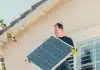 A Man in Black Shirt Standing on the Roof while Holding a Solar Panel