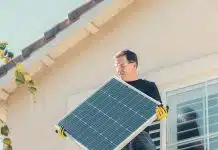 A Man in Black Shirt Standing on the Roof while Holding a Solar Panel